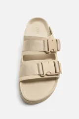 Sandalias planas color beige con doble tira ancha en el empeine y cierre con hebilla.