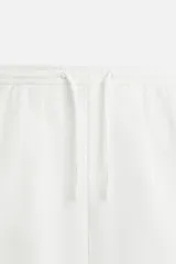 Bermuda color beige de corte recto, con cintura elástica ajustable con cordón, detalle de pliegues frontales, bolsillos laterales y bolsillos de vivo en la espalda.
