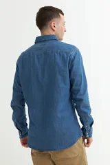 Camisa de denim azul con cuello clásico, cierre frontal con botones a presión, dos bolsillos con solapa abotonada en el pecho y mangas largas con puños abotonados.