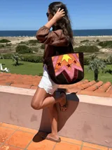 Bolso tote artesanal de cuero, reversible. Un lado presenta un diseño patchwork con figuras geométricas delimitadas por pasamanería en tonos tierra, naranja, amarillo y rosado. El reverso es liso.