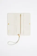 Billetera de cuero sintético color blanco con textura granulada. Tiene cierre con solapa y botón a presión, múltiples compartimentos internos para tarjetas y billetes, y un bolsillo con cierre para monedas. Incluye una correa de mano extraíble.