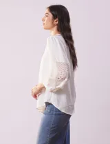 Blusa blanca de gasa con cuello a la base, escote en V y abertura central con botones. Tiene mangas 3/4 amplias con apliques de crochet y detalles plisados en la parte delantera.