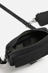 Bandolera negra de tela técnica con correa ajustable y extraíble. Tiene un compartimento principal con cierre de cremallera, un bolsillo interior con cremallera, un bolsillo frontal con solapa y hebilla de liberación rápida, y un monedero removible.