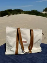 Bolso tipo tote de lona color crudo con asas de cuero marrón. Tiene un bolsillo interior y cierre superior.