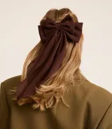 Accesorio para el pelo color marrón oscuro, con forma de lazo y caida de tejido. Modelo pinza.