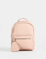 Mochila pequeña de cuerina color rosa pálido, con moño en relieve en el frente y llavero con forma de corazón trenzado. Tiene correas regulables, manija superior, cierre superior, bolsillo frontal con cierre y bolsillos laterales.