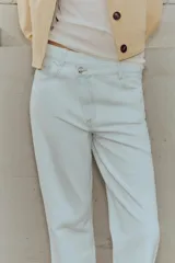 Jeans tobilleros de tiro alto color blanco, corte recto y bajo sin costura. Cierre frontal cruzado con cremallera y botones.