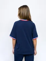 Remera blanca de corte clásico con cuello redondo y mangas cortas con puños tricolor (azul, blanco y rojo). Presenta el logo de Umbro bordado en el pecho izquierdo y el escudo del Club Nacional de Football bordado en el pecho derecho.