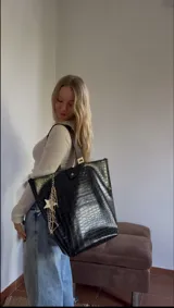 Bolso tote negro con textura símil cuero de cocodrilo, doble asa de hombro marrón y cierre con botón a presión. Incluye un charm colgante con una estrella blanca y cadenas plateadas.
