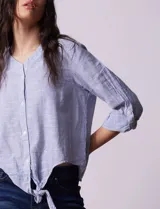 Blusa de algodón a rayas verticales azules y blancas, con cuello en V, mangas 3/4 y cierre frontal con botones. Tiene un nudo en la parte delantera.