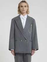 Blazer cruzado de lana gris con finas rayas blancas verticales (pinstripe). Presenta solapas anchas, doble botonadura con botones claros y un accesorio decorativo de cadena de perlas colgante prendido en la solapa izquierda.