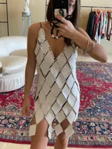 Vestido corto blanco, tipo túnica, confeccionado artesanalmente con cientos de rombos de cuero genuino perforados y unidos entre sí mediante engarces, creando un efecto de malla con aberturas. Presenta escote en V profundo y tirantes finos.