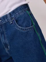 Bermuda de jean azul oscuro, corte holgado, con costuras en contraste color blanco y una franja lateral de ribete verde brillante.