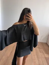 Conjunto negro brillante compuesto por un top corto con escote off-shoulder y mangas anchas, y una falda corta con lazo lateral.