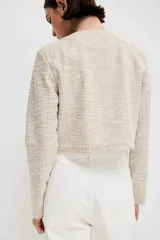 Saco corto de tweed color beige, con cuello redondo, cierre frontal con botones dorados y bolsillos delanteros decorativos.