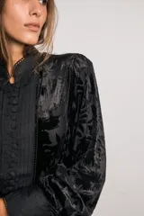 Camisa negra de terciopelo con motivos florales, cuello mao, mangas abullonadas, detalles plisados en el frente, botones forrados y bordes con puntilla.