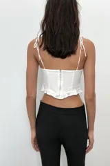 Top crop corsetero negro de popelín con escote recto, tirantes finos ajustables con lazada y bajo acabado con volante. Tiene forro interior a tono y cierre de cremallera en la espalda.