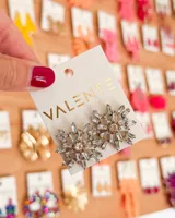 Aretes tipo 'stud' o botón, con forma de copo de nieve o estrella, compuestos por cristales transparentes facetados en forma de baguette dispuestos radialmente alrededor de un cristal central redondo de color champagne.