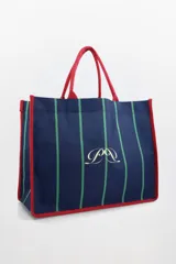 Bolso tote azul marino con rayas verticales verdes y ribetes rojos. Tiene asas de mano rojas y un logo dorado estampado en el centro. Incluye un pouch interno.
