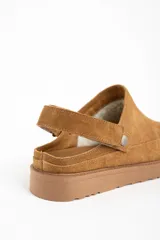 Zueco tipo mule de gamuza color camel, con forro interior de corderito visible en el borde. Presenta una tira ajustable con broche metálico en el lateral y suela de goma gruesa y dentada.