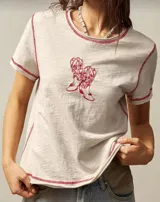 Remera blanca de algodón con cuello redondo y mangas cortas. Presenta un estampado de dos botas texanas rojas en el centro y costuras rojas a contraste en cuello, mangas y ruedo.