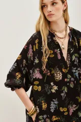 Vestido midi negro con estampado floral en tonos pastel y amarillos. Presenta escote pronunciado en V, mangas largas con puños anchos y detalles de bordado o pasamanería oscura en los bordes.