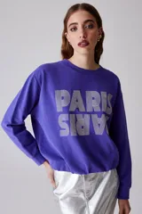 Buzo violeta de algodón con cuello redondo y mangas largas. Estampado frontal con la palabra "Paris" en color plateado.