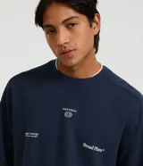 Remera de algodón azul marino, corte oversized, con cuello redondo y mangas cortas. Presenta un pequeño estampado en el pecho con texto y un logo circular, y un estampado más grande con el texto "Broad Plans" en la parte inferior derecha del frente.