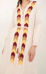 Saco color crema con bordado de flechas en tonos amarillo, marrón y rojo. Tiene mangas largas con detalle de puntilla y cinturón fino del mismo tejido.