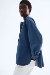 Blusa azul de denim con cuello redondo, tapeta con botones ocultos y mangas largas con puños abotonados.
