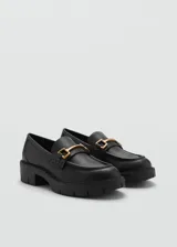 Zapato negro tipo loafer con suela gruesa y detalle de hebilla dorada.