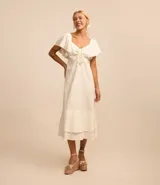 Vestido midi color crudo confeccionado en lino y viscosa, con escote cuadrado, mangas cortas con volados, detalle de lazo en el busto y falda con volados en el ruedo.