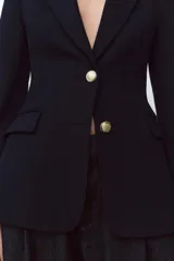 Blazer negra entallada con cuello solapa, manga larga con hombreras, bolsillos delanteros con solapa y cierre frontal con botones dorados.