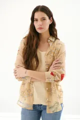 Camisa beige de manga larga con cuello clásico y cierre frontal con botones. Presenta bordados con motivos florales y geométricos en tonos azules, rojos y verdes.