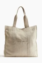 Bolso shopper de lona color beige con doble asa de hombro y cierre de botón a presión. Tiene un compartimento exterior y un compartimento interior pequeño, ambos abiertos.