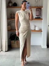 Vestido largo color crema, sin mangas, con cuello drapeado y fruncido lateral con detalle de argolla que genera una abertura frontal.