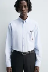 Camisa de algodón color celeste claro, corte regular fit, con cuello abotonado (button-down), manga larga con puño abotonado y bolsillo de parche en el pecho izquierdo.