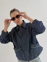 Campera de jean negra tipo bomber con cuello redondo y cierre frontal con botones metálicos. Presenta un parche rectangular con diseño gráfico en la espalda.