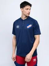 Remera de entrenamiento Umbro del Club Nacional de Football, color azul con detalles geométricos en mangas y hombros. Presenta cuello polo con botones, logo de Umbro y escudo del club en el pecho.
