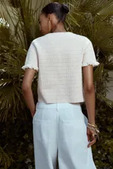 Chaqueta de punto color crema con cuello redondo, manga corta con volantes, bolsillos de plastrón y cierre frontal con botones metálicos.