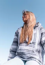 Buzo estilo mexicano con capucha, tejido en hilo grueso a rayas horizontales blancas y negras, con bolsillo tipo canguro y cordones ajustables en el cuello.