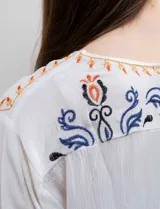Blusa blanca de rayón con escote en V y mangas largas. Presenta bordados florales en tonos naranja, azul y negro en la parte delantera y en las mangas.