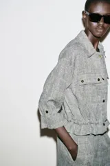 Chaqueta jaspeada color beige, con cuello solapa, manga tres cuartos con trabilla y botón, bolsillos delanteros de plastrón con solapa, bajo ajustable con cordones interiores y cierre frontal con botones a presión.