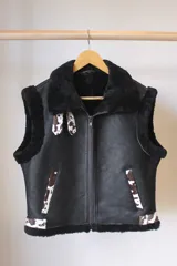 Chaleco negro de cuero con cuello de piel sintética y cierre frontal con cremallera. Presenta detalles de piel sintética con estampado animal en bolsillos y solapas.