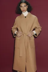 Abrigo largo color camel confeccionado en hilatura con lana. Tiene cuello clásico, cierre frontal con botones ocultos, bolsillos de parche y cinturón del mismo tejido.