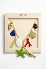 Colgante de fantasía con cadena dorada de eslabones irregulares y múltiples dijes temáticos marinos: caracoles dorados, concha azul, coral rojo, estrella de mar verde, disco dorado, óvalo blanco, dije azul con perla y pez dorado.