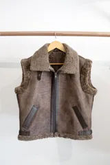 Chaleco negro de cuero con cuello y borde inferior de piel sintética negra. Cierre frontal con cremallera color cobre y detalles en cuero color tostado.