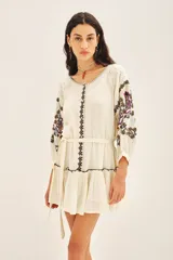 Vestido corto de bambula color celeste claro con bordados florales multicolor en el frente y mangas. Presenta escote redondo con cartera, cinturón de tela ajustable y volado en el ruedo.