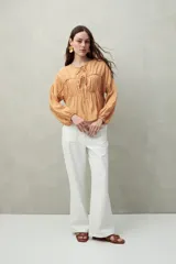 Blusa color beige de caída fluida con cuello redondo y lazos frontales. Diseño con cortes horizontales y frunces, mangas largas con puños elastizados.