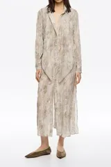 Vestido largo de corte recto confeccionado en tejido plisado con estampado de piel de serpiente en tonos beige y marrón. Presenta botones frontales, cuello camisero, mangas largas con puño y botón, y maxilazos en el escote delantero.